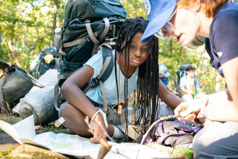 Two people are looking at a map in a wooded area. One person is wearing a large backpack and has long braids. The other person is wearing a blue hat and glasses. They appear to be planning a route or checking their location. There are other backpacks and camping gear nearby, suggesting they are on a hiking or camping trip.